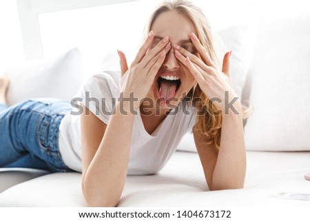 Beautiful young woman relaxing on a couch at home, showing tongue, covering eyes