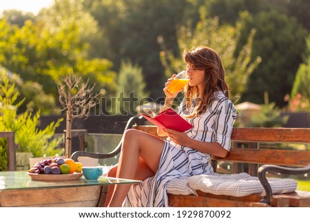 Beautiful young woman relaxing on a sunny summer morning in her backyard, having breakfast, drinking orange juice and reading a book outdoors
