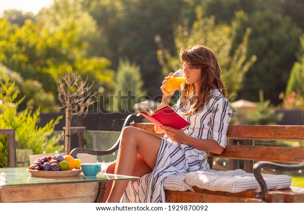 Beautiful young woman relaxing on a sunny summer\
morning in her backyard, having breakfast, drinking orange juice\
and reading a book\
outdoors