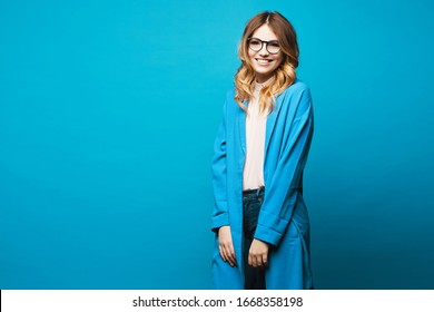 Beautiful young woman in round glasses, beige blouse and trendy blue coat isolated at blue background. Model girl in blouse, blue coat and modish eyewear over blue background. Copy space. Spring look