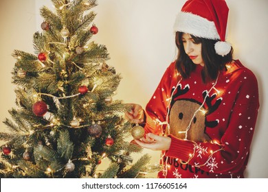beautiful young woman in santa hat hand holding golden glitter ornament, decorating  christmas tree with lights, preparation for holidays in festive room. decor for winter holidays