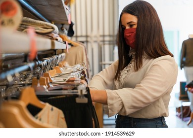 Beautiful Young Woman In Shop Looks At Clothes To Buy Wearing Protective Face Mask During Coronavirus Pandemic Covid-19 - Shop Assistant Arranges Dress