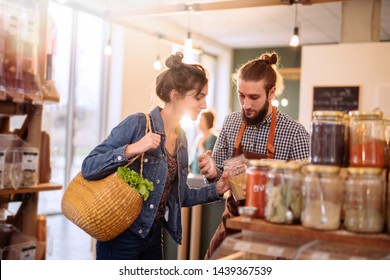 Beautiful Young Woman Shopping In A Bulk Food Store. The Seller Advises Her To Buy Organic Spices In Jars.