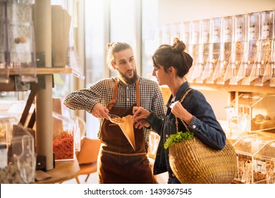 Beautiful Young Woman Shopping In A Bulk Food Store. The Seller Advises Her On Her Purchases Of Organic Food