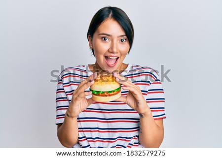Beautiful young woman with short hair eating hamburger celebrating crazy and amazed for success with open eyes screaming excited. 