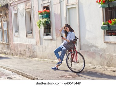 Beautiful Young Woman Siting On Bicycle In Old Town And Looking At Mobile Phone.