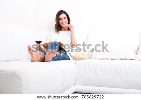 A beautiful young woman siting on a couch reading a book drinking wine and eating popcorn