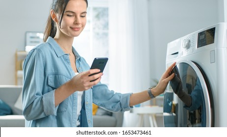 Beautiful Young Woman Sits on Her Knees Next to the Washing Machine. She Loaded the Washer with Dirty Laundry and Configured the Wash With Her Smartphone. Shot in Living Room with Modern Interior. - Powered by Shutterstock