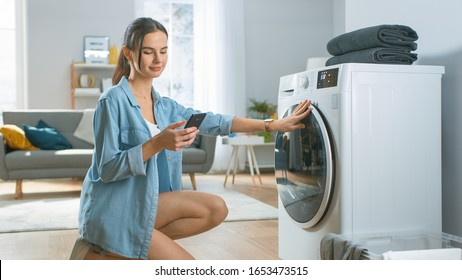 Beautiful Young Woman Sits on Her Knees Next to the Washing Machine. She Loaded the Washer with Dirty Laundry While Using Her Smartphone. Shot in Living Room with Modern Interior. - Powered by Shutterstock