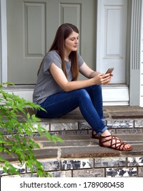 Beautiful young woman sits on the steps of her home holding her cell phone.  She has her eyes closed listening to her music. - Shutterstock ID 1789080458