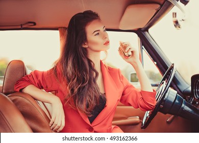Beautiful young woman sitting behind the wheel in retro automobile using a perfume