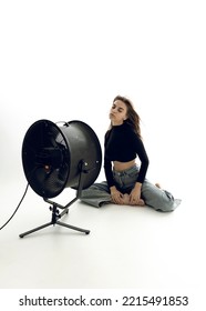 A Beautiful Young Woman Is Sitting In A Bright Room On The Floor Cooling Down Under A Fan	
