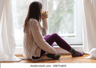 Beautiful young woman sitting by the window having hot drink