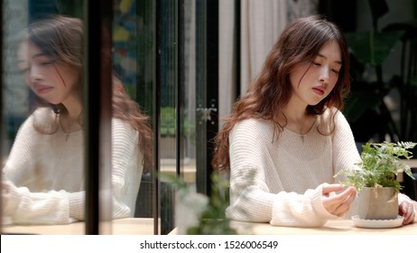 Beautiful young woman sitting in cafe and looking at the green plant on table in sunny day, with her mirror image in glass windows, lonely expression and vintage color. - Powered by Shutterstock