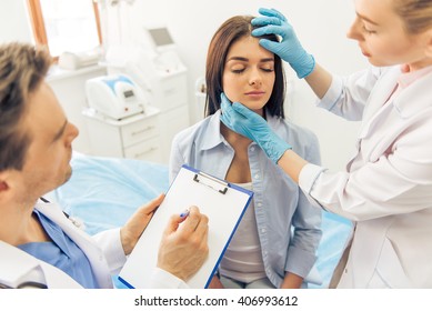 Beautiful young woman is sitting with closed eyes in doctors office, two doctors are examining her face and making notes