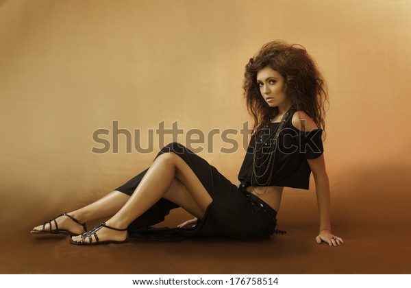 \
Beautiful young woman sitting down in a sexy black outfit with wild\
hair and gladiator sandals looking over her\
shoulder