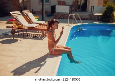 Beautiful young woman sitting at the edge of a swimming pool, relaxing on summer vacation, sunbathing and eating ice cream
