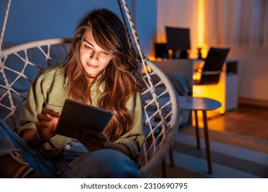 Beautiful young woman sitting in hanging chair reading an ebook and relaxing at home