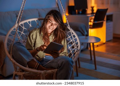 Beautiful young woman sitting in hanging chair surfing the net using tablet computer while relaxing at home