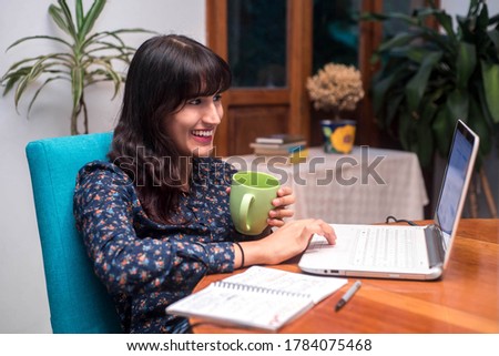 beautiful young woman sitting at home infront of the computer while holding a cup of coffee