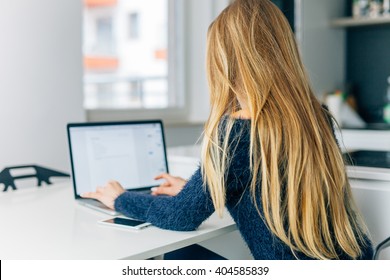 Beautiful Young Woman Sitting In The Kitchen With Laptop. Back View