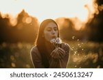 Beautiful Young Woman sitting on the field in green grass and blowing dandelion during a golden hour