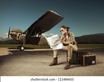 Beautiful young woman sitting on a suitcase, looks at a map. Airplane in the background