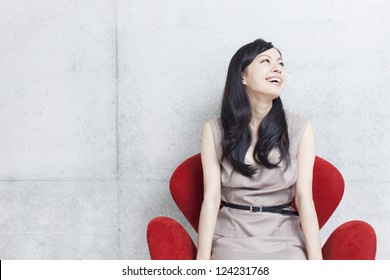 Beautiful Young Woman Sitting On Modern Chair Against Concrete Wall.