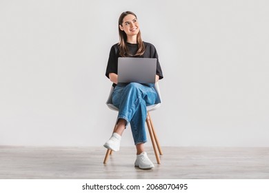 Beautiful Young Woman Sitting On Chair, Working On Laptop Against White Studio Wall, Full Length. Dreamy Millennial Lady Using Portable Pc For Work, Entertainment Or Online Studies