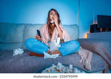 Beautiful young woman sitting on couch covered with blanket late at night, sad while watching drama movie on TV, crying and wiping tears with paper tissues - Powered by Shutterstock