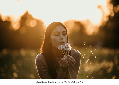 Beautiful Young Woman sitting on the field in green grass and blowing dandelion during a golden hour - Powered by Shutterstock