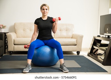 Beautiful Young Woman Sitting On A Stability Ball And Doing Bicep Curls With Dumbbells At Home