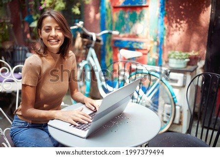 beautiful young woman sitting in outdoor cafe with her laptop on table, smiling looking at camera. eye contact