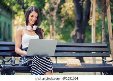 Beautiful young woman sitting in public park chatting over laptop computer