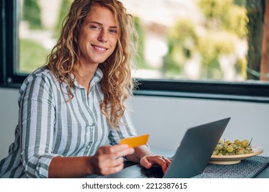 Beautiful Young Woman Sitting At The Table In The Kitchen At Home Using Laptop And A Credit Card. Blonde Female Paying Bill Or Online Shopping. Looking Into The Camera. Copy Space.