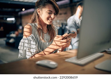 Beautiful young woman sitting at table in front of desktop computer using smartphone, feeling joyful. Business, student, office job concept. - Powered by Shutterstock