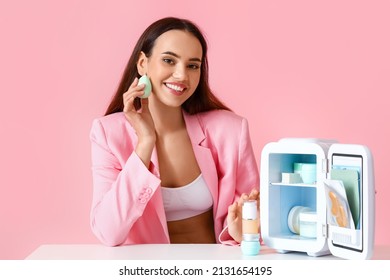 Beautiful young woman with small refrigerator and cosmetic products sitting at table on color background