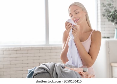 Beautiful young woman smelling clean clothes and smiling while doing laundry at home. Caucasian housewife touching soft clothes after washing machine.