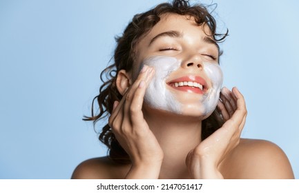 Beautiful young woman smiling, applying facial cream, scrub or cleansing product on her face with joy and delight, standing over blue background