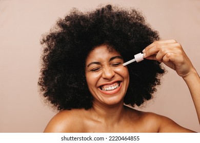 Beautiful Young Woman Smiling Cheerfully While Applying Face Serum Using A Dropper. Happy Woman With Afro Hair Treating Her Skin With A Nourishing Beauty Product.