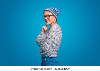 Beautiful young woman smiling gently keeping hand under chin looking at camera with dreamy expression wearing knitted hat isolated over blue background.