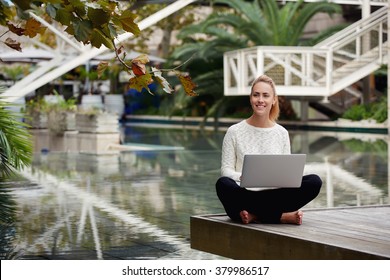 Beautiful Young Woman Smiling To Someone During Work On Laptop Computer Outside In Autumn Park, Cheerful Hipster Girl Holding Open Net-book On Knees While Sitting On A Wooden Pier In Warm Fall Day