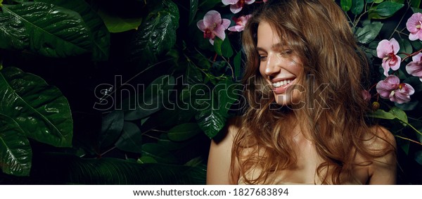 Beautiful young woman with\
smooth tanned skin laughs cheerfully while standing against the\
background of exotic plants and flowers. Natural cosmetics, skin\
care.