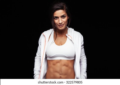 Beautiful Young Woman In Sports Clothing Smiling While Standing Against Black Background. Confident Female Fitness Model With Muscular Abs.