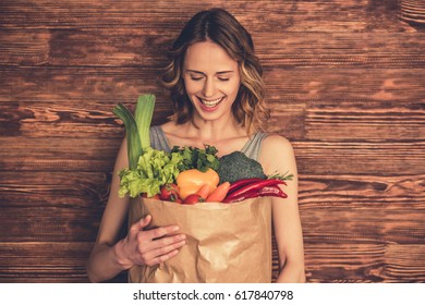 Beautiful Young Woman In Sportswear Is Holding A Shopping Bag Full Of Healthy Food, Looking At Camera And Smiling, On Wooden Background