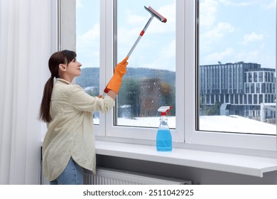 Beautiful young woman with squeegee tool cleaning window indoors - Powered by Shutterstock