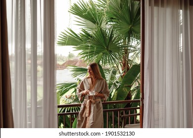 Beautiful Young Woman Stand  On Balcony Terrace Of Jungle Villa With Morning Coffee, Wearing Bathrobe. Vacation On Tropical Island