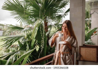 Beautiful Young Woman Stand  On Balcony Terrace Of Jungle Villa With Morning Coffee, Wearing Bathrobe. Vacation On Tropical Island