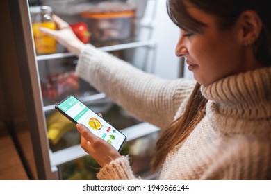Beautiful young woman standing next to an opened refrigerator door, holding a smart phone and ordering fresh fruit and vegetables online for home delivery