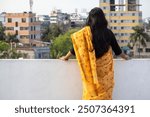 Beautiful young woman standing on roof of house in yellow saree looking away. Saree is traditional and popular garment for women in Bangladesh. Backside view.